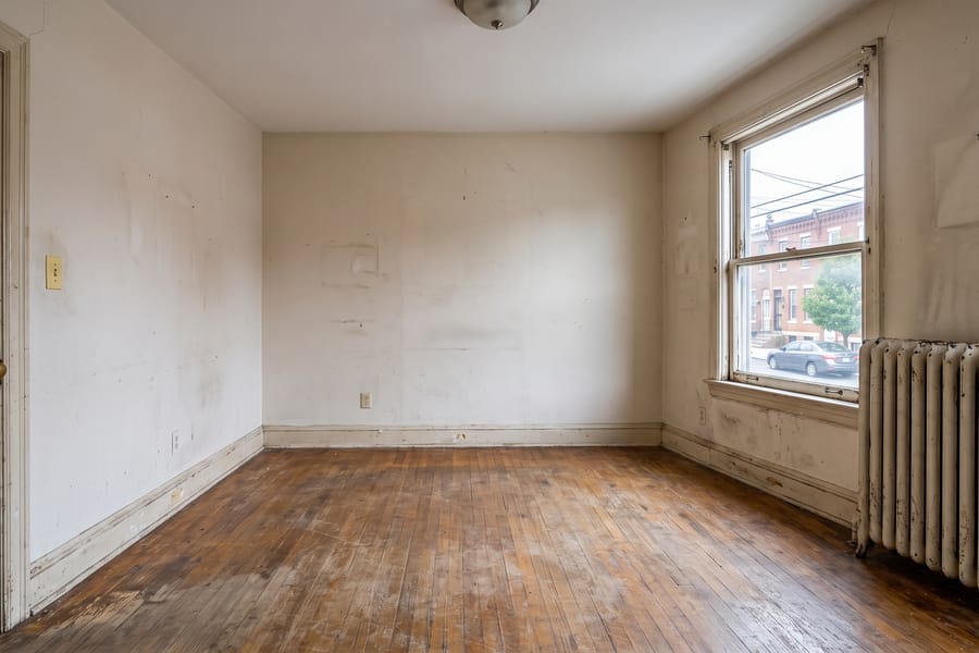 An empty, tired rental room in a Baltimore rowhome with worn wood floors, scuffed walls, and an old radiator.